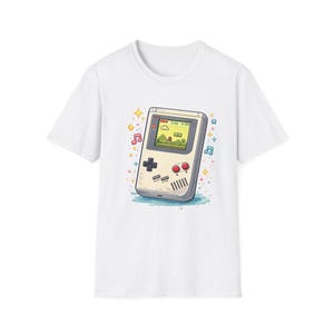 May include: White t-shirt featuring a cartoon illustration of a retro handheld video game console. The game console is light gray with a green screen displaying a pixelated landscape. Musical notes and stars surround the console.