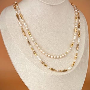 May include: Two layered necklace displayed on a mannequin. The necklaces feature white pearls and gold-colored beads. The pearls are of varying sizes and shapes. The gold beads are small and interspersed throughout the necklace.