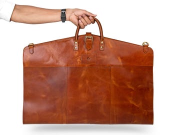 Puede incluir: Una funda de ropa de cuero marrón con asa y cierre de hebilla. La bolsa tiene forma rectangular con la parte superior curvada y la sostiene una mano. La bolsa tiene un pequeño logotipo en la parte delantera.