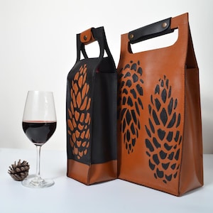 May include: Two leather wine bottle carriers. One is black with a brown pinecone design, and the other is brown with a black pinecone design. A wine glass with red wine and a pinecone are in the foreground.