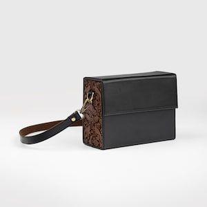 Könnte beinhalten: Schwarze rechteckige Handtasche mit Klappenverschluss. Eine Seite hat ein braunes Blumenmuster, während die anderen Seiten einfarbig schwarz sind. Sie hat einen kurzen, schwarzen Griff und goldfarbene Beschläge.