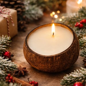May include: A lit candle in a coconut shell, surrounded by festive decorations. The candle has a white wax and a single flame. The decorations include pine needles, red berries, and a gift box. The scene is set on a wooden surface.