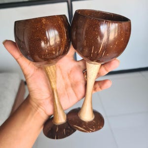 May include: Two wine glasses crafted from coconut shells. The dark brown bowls sit on light brown wooden stems and bases. These glasses have a natural, rustic appearance, perfect for a tropical theme.