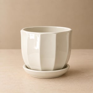 Peut inclure: Un pot de fleurs en céramique blanc cassé brillant avec un design extérieur facetté à plusieurs côtés. Le pot présente une large ouverture sur le dessus et repose solidement sur une soucoupe en céramique assortie, formant un ensemble cohérent.