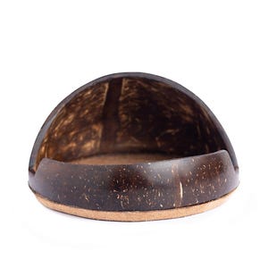 May include: A dark brown, semi-circular soap dish made from a coconut shell. The dish has a natural, textured surface and a light brown base. The soap dish is designed to hold a bar of soap. The dimensions are in centimetres.