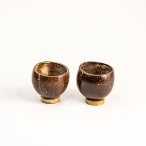 May include: Two handcrafted coconut shell cups with a natural brown finish. Each cup has a rounded body and a slightly angled opening. The cups are set on small, circular, light-colored bases, and are ideal for serving drinks or as decorative items.