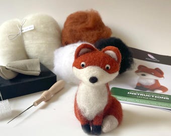 May include: A needle felting kit featuring a small, orange and white fox figurine. The kit includes wool in various colors, needles, a foam pad, and instructions. The finished fox is approximately 4 inches tall.