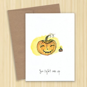 May include: A watercolor illustration of a smiling pumpkin with a lit candle next to it. The text "you light me up" is written below the pumpkin.