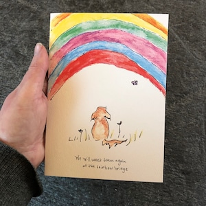 May include: A watercolor illustration of a rainbow with a small butterfly above it. A brown dog with a long tail is sitting under the rainbow with a small plant on either side of it. The text "We will meet them again at the rainbow bridge" is written below the dog.