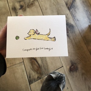 May include: A white card with a hand-drawn illustration of a yellow puppy with a pink bow running towards a green ball. The card says "Congrats on your fur baby!"