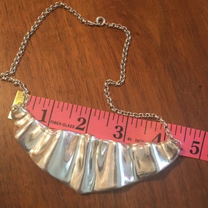 May include: A silver necklace featuring a wide, curved pendant with a pleated texture. The chain is a simple link design. A pink measuring tape is placed next to the necklace. The pendant is approximately 7.6 cm wide.