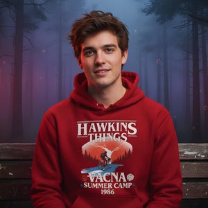 May include: A red hoodie with the text "Hawkins Things" and a graphic of a person on a bicycle in front of a mountain and the text "Vacna Summer Camp 1986". The hoodie has a hood and a drawstring.