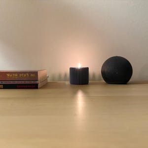 Könnte beinhalten: Eine brennende graue Kerze in einem gerippten Halter, eine dunkle Kugel und zwei Bücher liegen auf einer hellen Holzoberfläche. Die Kerzenflamme leuchtet hell. Die Bücher tragen die Titel "The Last Bear" und "Bad Castro".