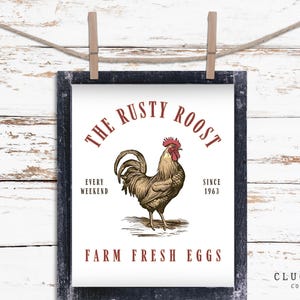 Op de afbeelding: Een ingelijste print met een rustiek ontwerp met een illustratie van een haan. De tekst luidt "The Rusty Roost" boven de haan, met "Farm Fresh Eggs" eronder. De print wordt vastgehouden door houten wasknijpers.