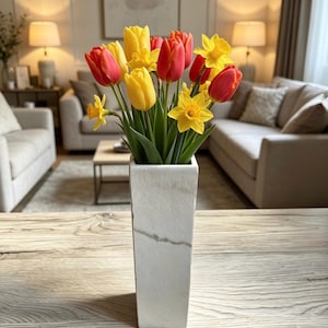Op de afbeelding: Een witte rechthoekige vaas gevuld met een levendig arrangement van rode, gele en oranje tulpen en narcissen. De vaas staat op een lichtgekleurde houten tafel, met een vage woonkamer op de achtergrond.