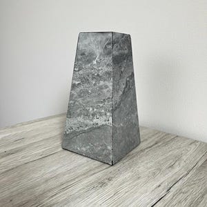 Könnte beinhalten: Eine graue, marmoriert strukturierte, trapezförmige Skulptur. Der Stein hat ein gesprenkeltes Muster aus hellem und dunklem Grau. Die Skulptur steht auf einer hellgrauen Holzoberfläche. Die Skulptur ist ein dekoratives Objekt.