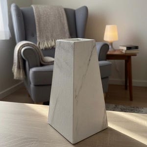 Può includere: Un vaso geometrico bianco con un motivo simile al marmo. Il vaso ha una parte superiore quadrata e una forma piramidale affusolata. Si trova su una superficie di legno chiaro, con uno sfondo morbido con una sedia e una lampada.