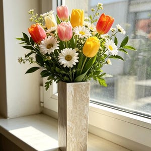 May include: A floral arrangement of colorful tulips and white daisies in a tall, rectangular vase. The tulips are in shades of orange, yellow, and pink. The vase is a neutral beige color and sits on a windowsill.