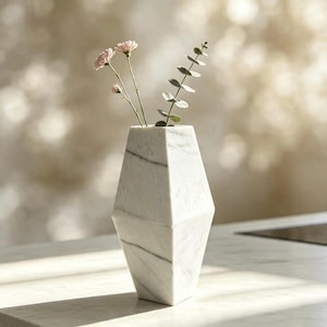 Op de afbeelding: Een witte marmeren vaas met een geometrische, gefacetteerde vorm en grijze aders. De vaas bevat drie lichtroze bloemen op dunne stelen en een groene eucalyptus-tak met kleine, ronde blaadjes die uit de bovenste opening steken.