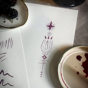 Op de afbeelding: Een stilleven met een wit bord met bramen, een pot bramenjam en een paarse tekening met een ster en geometrische vormen. De tekening staat op wit papier, op een grijze ondergrond.