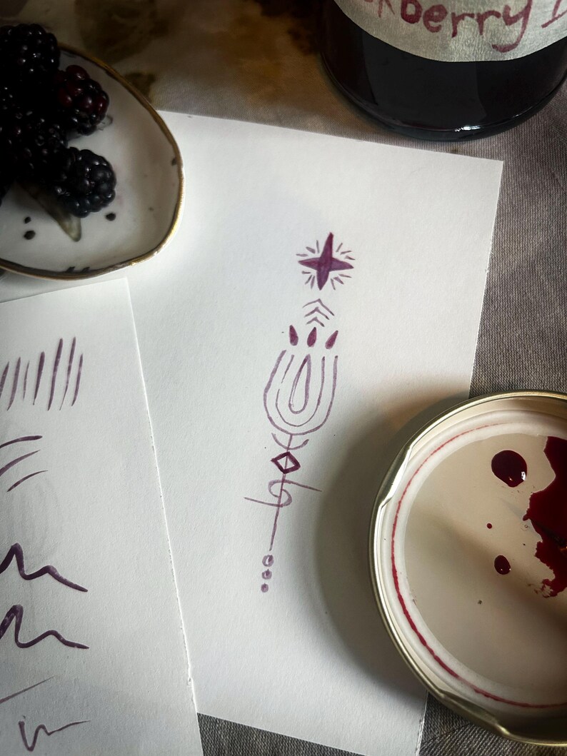 Op de afbeelding: Een stilleven met een wit bord met bramen, een pot bramenjam en een paarse tekening met een ster en geometrische vormen. De tekening staat op wit papier, op een grijze ondergrond.