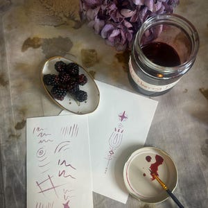 Op de afbeelding: Een stilleven met bramen, een pot bramenjam en aquarel schetsen. De pot is gelabeld met "bramenjam". Een kleine penseel ligt op een deksel met rode verf. Een tros gedroogde paarse bloemen staat op de achtergrond.