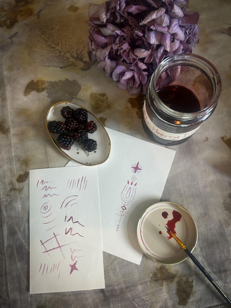 Op de afbeelding: Een stilleven met bramen, een pot bramenjam en aquarel schetsen. De pot is gelabeld met "bramenjam". Een kleine penseel ligt op een deksel met rode verf. Een tros gedroogde paarse bloemen staat op de achtergrond.