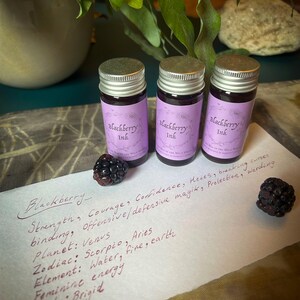 Op de afbeelding: Drie kleine glazen flesjes Blackberry Ink met zilveren deksels en paarse etiketten. De etiketten bevatten de tekst "Blackberry Ink". Twee bramen en handgeschreven tekst zijn ook zichtbaar.