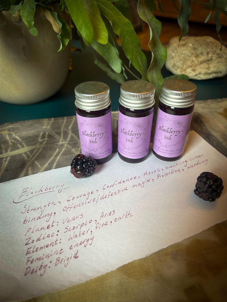 Op de afbeelding: Drie kleine glazen flesjes Blackberry Ink met zilveren deksels en paarse etiketten. De etiketten bevatten de tekst "Blackberry Ink". Twee bramen en handgeschreven tekst zijn ook zichtbaar.