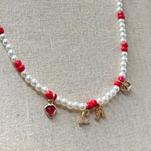 Könnte beinhalten: Eine zarte Halskette mit weißen Kunstperlen und leuchtend roten Perlen. Die Halskette enthält einen kleinen roten Herzanhänger und goldfarbene Initialen-Anhänger. Die Halskette wird vor einem neutralen Hintergrund präsentiert.