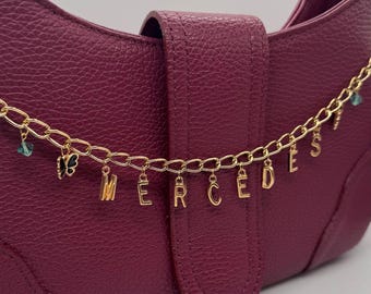 May include: A deep burgundy handbag with a gold chain strap. The chain features small charms and the name "MERCEDES" spelled out in gold letters. The bag has a textured surface and a flap closure.