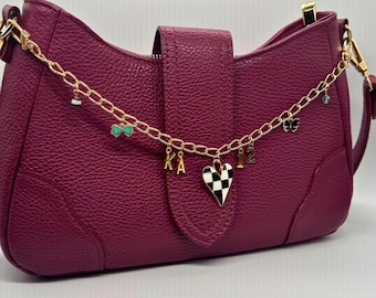 May include: A deep magenta textured shoulder bag featuring a gold-tone chain strap adorned with various charms, including the letters KA, the number 12, a black and white checkered heart, a small butterfly, and a turquoise bow accent.