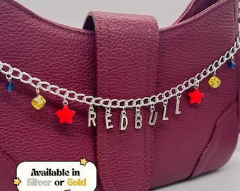 May include: A burgundy handbag with a silver chain. The chain has red star charms, yellow dice charms, and the word "REDBULL" spelled out in silver letters. The text "Available in Silver or Gold" is in a white box.