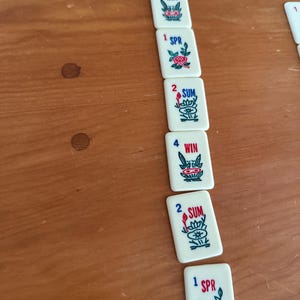 K&ouml;nnte beinhalten: Eine Ansammlung wei&szlig;er Mahjong-Steine mit roter und blauer Schrift und floralen Designs. Die Steine zeigen Zahlen und Saisonnamen wie AUT, WIN, SPR und SUM. Die Steine liegen auf einer Holzoberfl&auml;che.