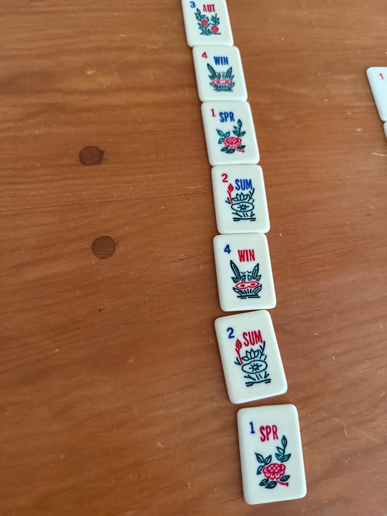 K&ouml;nnte beinhalten: Eine Ansammlung wei&szlig;er Mahjong-Steine mit roter und blauer Schrift und floralen Designs. Die Steine zeigen Zahlen und Saisonnamen wie AUT, WIN, SPR und SUM. Die Steine liegen auf einer Holzoberfl&auml;che.