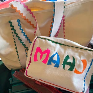 K&ouml;nnte beinhalten: Cremefarbene Tragetasche und passende Tasche. Die Tragetasche hat wei&szlig;e Tr&auml;ger und einen farbenfrohen, gewellten Besatz in Pink, Orange, Blau und Gr&uuml;n. Die Tasche zeigt das Wort "MAHU" in leuchtendem Pink, Blau, Gr&uuml;n und Orange.