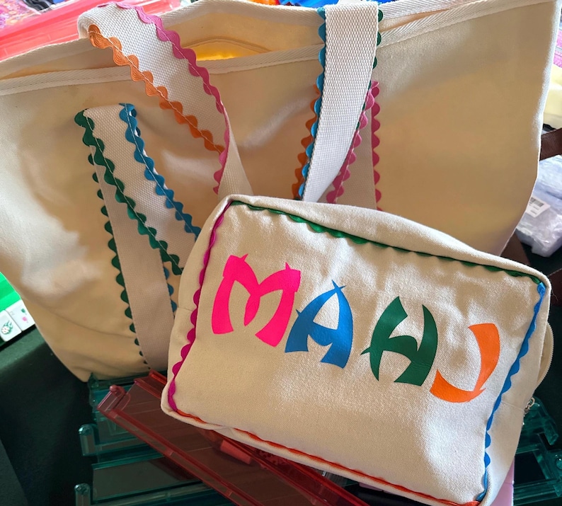 K&ouml;nnte beinhalten: Cremefarbene Tragetasche und passende Tasche. Die Tragetasche hat wei&szlig;e Tr&auml;ger und einen farbenfrohen, gewellten Besatz in Pink, Orange, Blau und Gr&uuml;n. Die Tasche zeigt das Wort "MAHU" in leuchtendem Pink, Blau, Gr&uuml;n und Orange.
