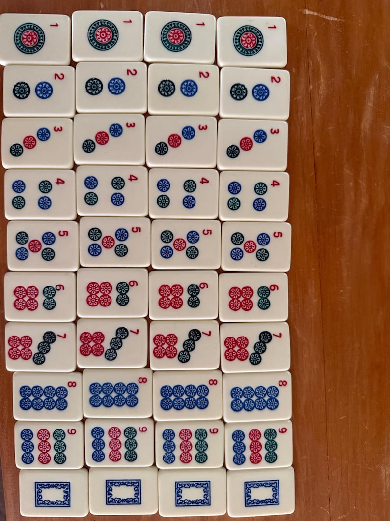K&ouml;nnte beinhalten: Eine Sammlung rechteckiger Mahjong-Steine. Jeder Stein ist wei&szlig; mit schwarzen, roten und blauen Symbolen und Zahlen. Die Steine sind auf einer Holzoberfl&auml;che angeordnet und zeigen verschiedene Kombinationen aus Punkten und Zeichen.