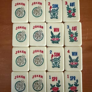 K&ouml;nnte beinhalten: Eine Sammlung elfenbeinfarbener Mahjong-Steine. Die Steine zeigen rote, gr&uuml;ne und blaue Symbole und Texte, darunter "JOKER", Zahlen und saisonale Begriffe. Die Steine sind auf einer Holzoberfl&auml;che angeordnet.