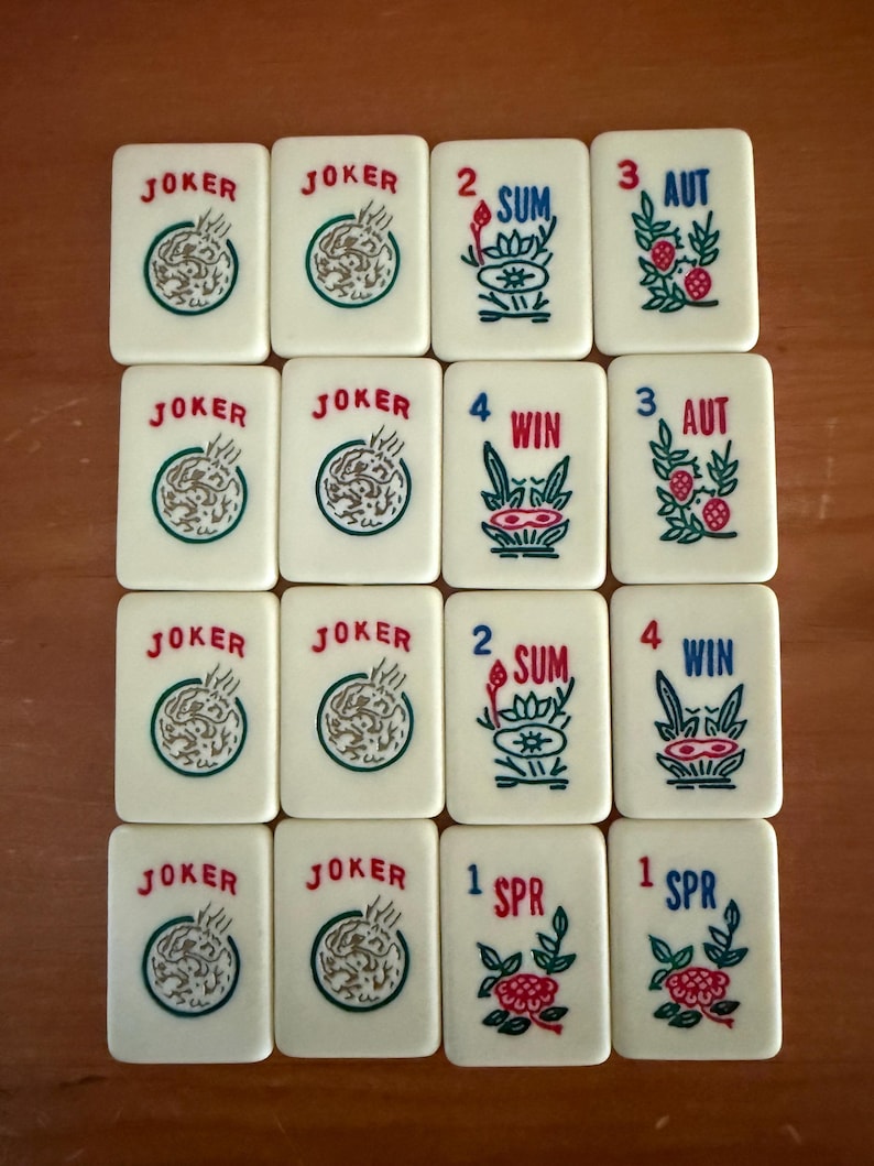 K&ouml;nnte beinhalten: Eine Sammlung elfenbeinfarbener Mahjong-Steine. Die Steine zeigen rote, gr&uuml;ne und blaue Symbole und Texte, darunter "JOKER", Zahlen und saisonale Begriffe. Die Steine sind auf einer Holzoberfl&auml;che angeordnet.