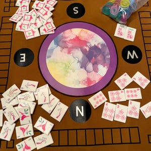 May include: A Mahjong game set with white tiles featuring various symbols and numbers, scattered on a brown mat. The mat has a central circular design with colorful abstract art and directional markers.