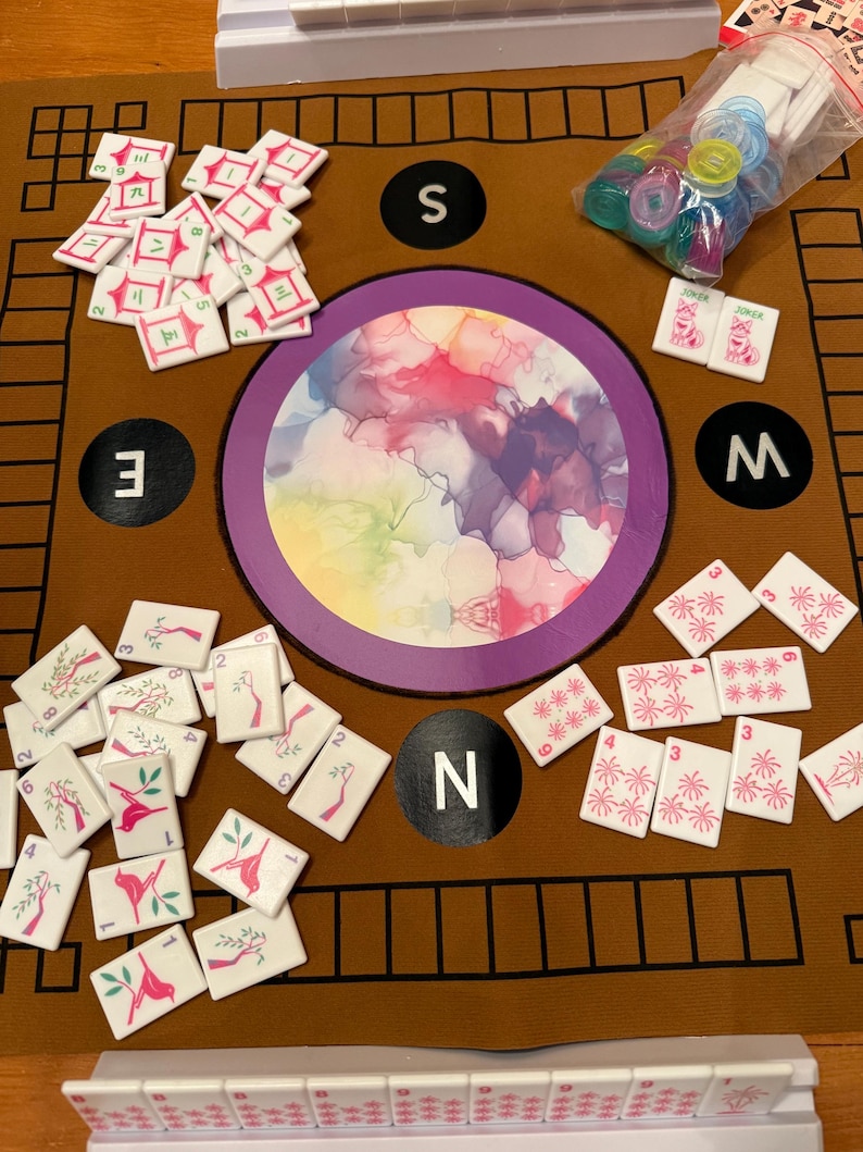 May include: A Mahjong game set with white tiles featuring various symbols and numbers, scattered on a brown mat. The mat has a central circular design with colorful abstract art and directional markers.