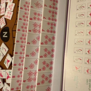 May include: A close-up of a Mahjong set with white tiles featuring red floral designs and numbers. The tiles are arranged in rows, with some scattered around. A game guide is visible on the right.