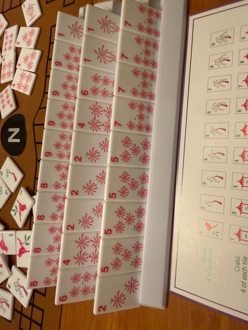 May include: A close-up of a Mahjong set with white tiles featuring red floral designs and numbers. The tiles are arranged in rows, with some scattered around. A game guide is visible on the right.