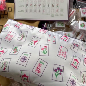 May include: A white cosmetic bag with a repeating pattern of Mahjong tiles in pink and green. The tiles feature various symbols, numbers, and the word "JOKER". A Mahjong tile set and game instructions are also visible.
