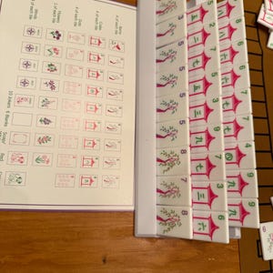 May include: A Mahjong set with white tiles featuring red and green symbols and numbers. The set includes a booklet with illustrations of the tiles. The tiles are arranged on a wooden surface.