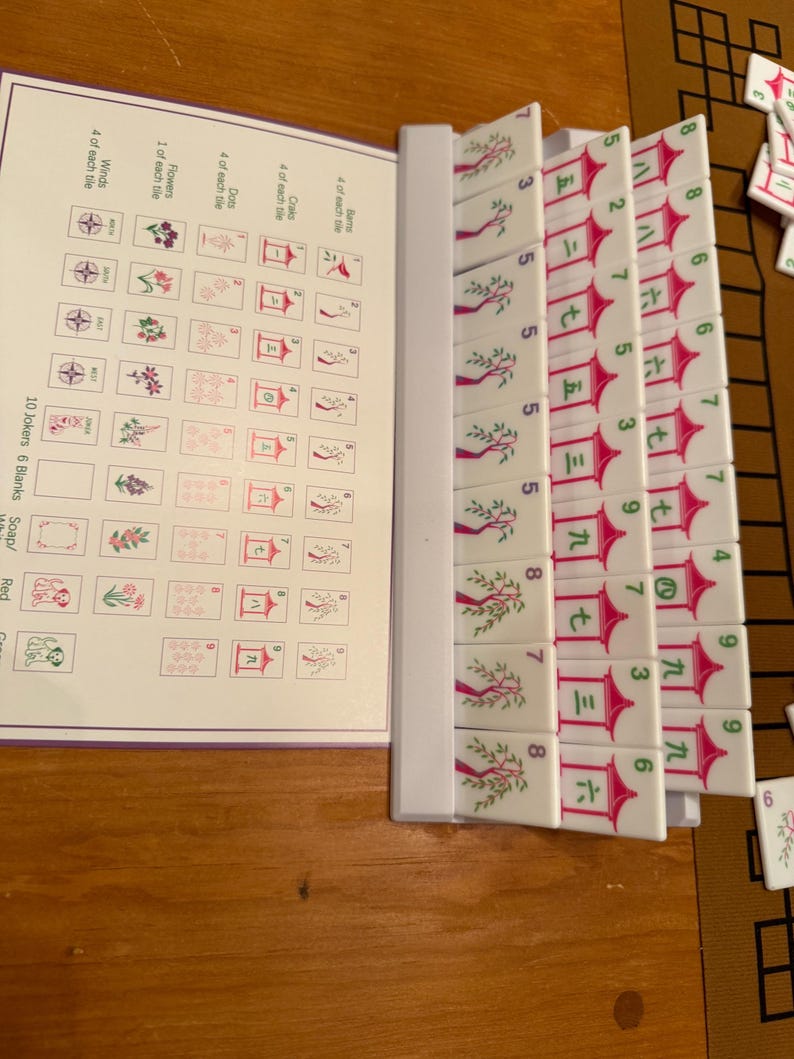 May include: A Mahjong set with white tiles featuring red and green symbols and numbers. The set includes a booklet with illustrations of the tiles. The tiles are arranged on a wooden surface.