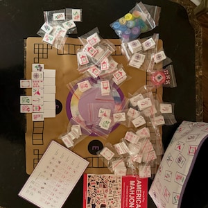 May include: An American Mahjong set with tiles, dice, and a book titled "American Mahjong". The tiles are white with red and green symbols. The set includes a purple and white carrying case and a game board.