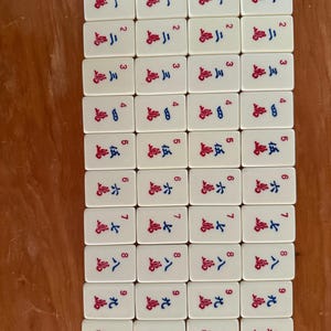 K&ouml;nnte beinhalten: Eine Sammlung rechteckiger Mahjong-Steine. Jeder Stein ist wei&szlig; mit roten und blauen chinesischen Schriftzeichen und Zahlen. Die Steine sind in einem Rastermuster auf einer Holzoberfl&auml;che angeordnet. Die Steine sind von 1 bis 9 nummeriert.