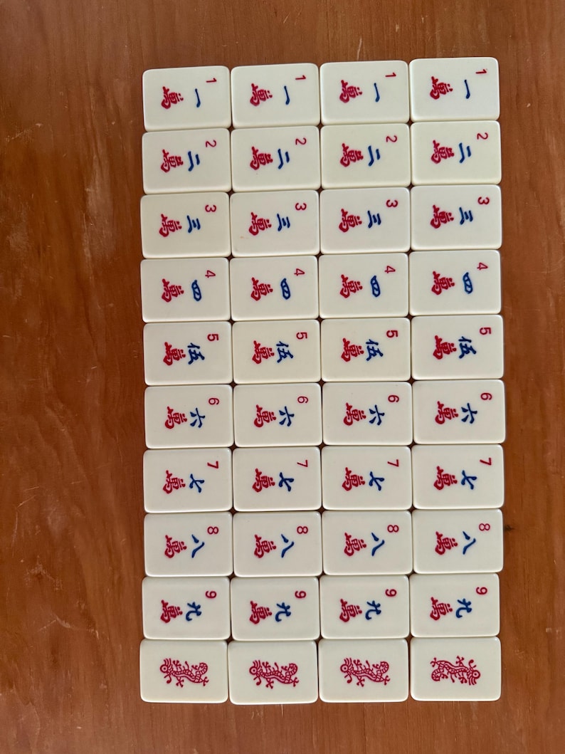 K&ouml;nnte beinhalten: Eine Sammlung rechteckiger Mahjong-Steine. Jeder Stein ist wei&szlig; mit roten und blauen chinesischen Schriftzeichen und Zahlen. Die Steine sind in einem Rastermuster auf einer Holzoberfl&auml;che angeordnet. Die Steine sind von 1 bis 9 nummeriert.